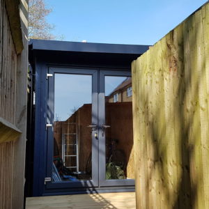 Modern garden office with glass double doors, painted in dark blue, surrounded by wooden fencing. Inside, there are visible items including a ladder and a bicycle. The blue sky is visible above. - a room in the garden