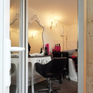 A well-lit hair salon with an ornate mirror and chandelier. A black styling chair is positioned in front of a white vanity table holding various hair products. In the background, a hair washing station with a black sink and a rolling cart can be seen. - a room in the garden