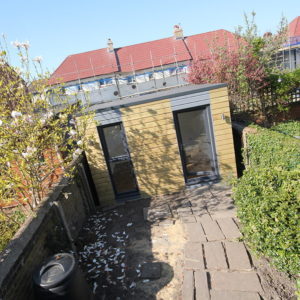 A small, modern garden shed with a gray roof and wooden exterior stands at the end of a narrow pathway made of stone slabs. It is surrounded by greenery, including budding trees and a trimmed hedge. In the background, a row of houses with red roofs is visible. - a room in the garden