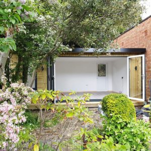 A bright, modern garden room designed by a top garden room designer, with an open front surrounded by lush greenery. The structure boasts a wooden exterior, large glass windows, and a small door at the back. A variety of plants and blooming flowers adorn the foreground. - a room in the garden