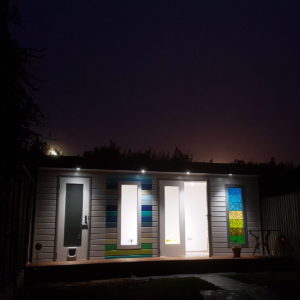A modern, small, white gallery with three illuminated doors at night. The doors feature vertical rectangular windows; one has multicolored blue and green panes. Exterior lights shine above, and the moon is partially visible in the hazy sky. A potted plant sits outside. - a room in the garden