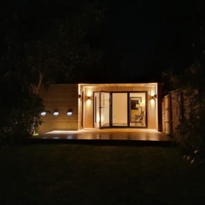 A modern, single-story house is illuminated at night. The exterior features sleek wooden paneling and large glass doors. Lights on each side of the entrance and three round lights on the left wall cast a warm glow, highlighting the minimalistic design of the porch. - a room in the garden