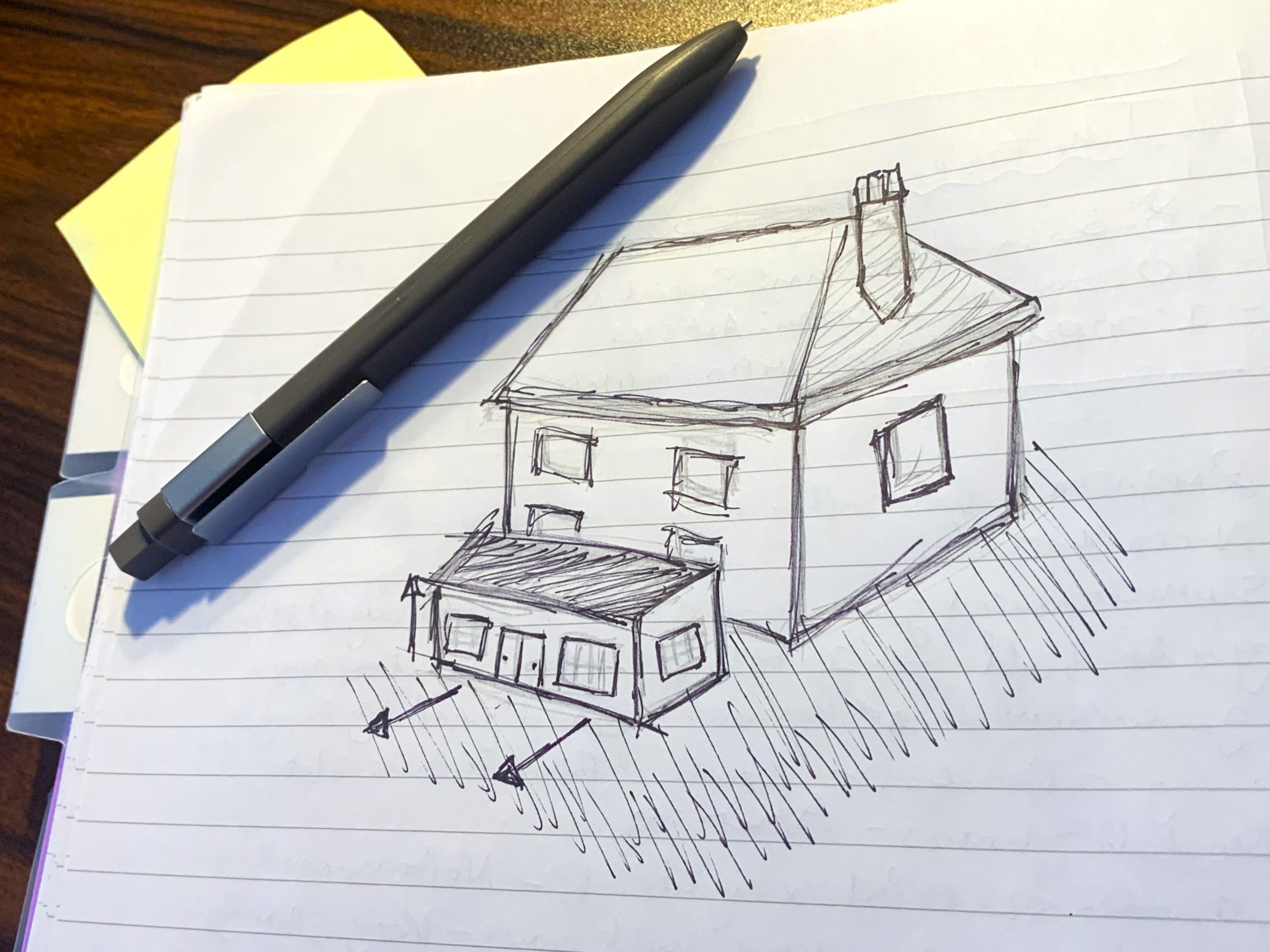 A black pen rests on a lined notebook page containing a sketch of a house with a chimney and an attached garage, meticulously annotated to meet building regulations. Various notes and papers are visible in the background. The sketch includes directional arrows and crosshatching, indicating shading and structural details. - a room in the garden