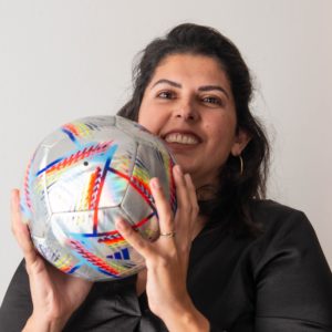 A person with dark hair and a black top is smiling, holding a colorful soccer ball close to their face with both hands. The background is a plain, light-colored wall, adding emphasis to their joyful expression about the moment. - a room in the garden
