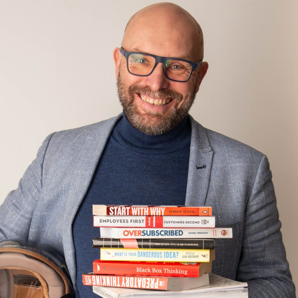 A smiling bald man with glasses holds a stack of books in his arm. He wears a blue blazer over a blue turtleneck. The titles of the books, including “Start With Why,” “Oversubscribed,” and “Predatory Thinking,” suggest he’s about self-improvement. In his other hand, he carries a helmet. – a room in the garden A smiling bald man with glasses holds a stack of books in his arm. He wears a blue blazer over a blue turtleneck. The titles of the books, including "Start With Why," "Oversubscribed," and "Predatory Thinking," suggest he's about self-improvement. In his other hand, he carries a helmet. - a room in the garden