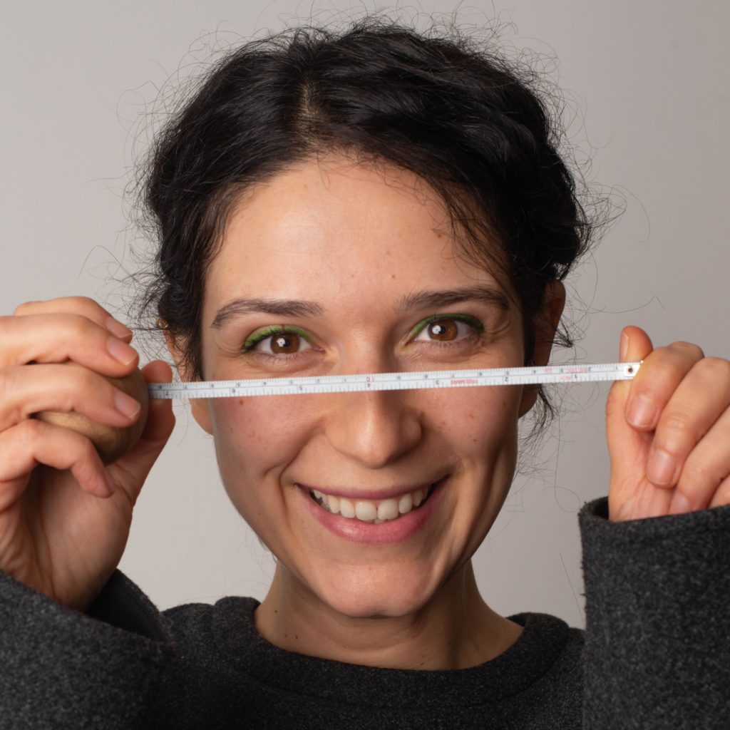 A woman with dark hair smiles and holds a small ruler horizontally in front of her face, level with her eyes. She is wearing a dark top and standing against a plain background, looking about as happy as can be. – a room in the garden A woman with dark hair smiles and holds a small ruler horizontally in front of her face, level with her eyes. She is wearing a dark top and standing against a plain background, looking about as happy as can be. - a room in the garden