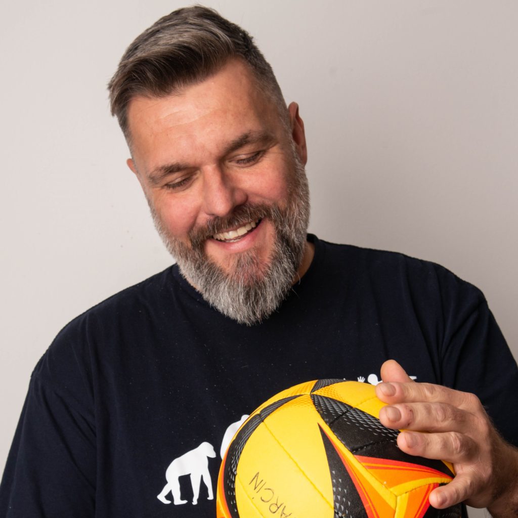 A bearded man wearing a navy shirt with a white gorilla graphic smiles as he looks at and holds a yellow and black soccer ball, clearly passionate about the sport. – a room in the garden A bearded man wearing a navy shirt with a white gorilla graphic smiles as he looks at and holds a yellow and black soccer ball, clearly passionate about the sport. - a room in the garden
