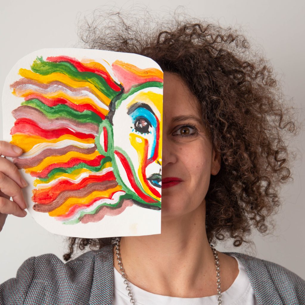 A woman with curly hair is holding a colorful painting of a face about in front of her own, aligning the painting’s eye and cheek with her own. The painting features wavy lines in red, yellow, and green extending from the face like hair. – a room in the garden A woman with curly hair is holding a colorful painting of a face about in front of her own, aligning the painting's eye and cheek with her own. The painting features wavy lines in red, yellow, and green extending from the face like hair. - a room in the garden