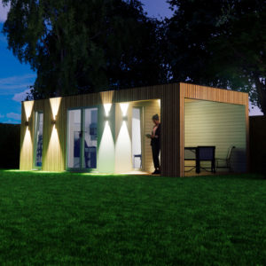 A modern, small house with wooden exterior walls and large glass doors is illuminated at night by bright outdoor lights. The grass lawn in front is vibrant green. A person stands near one of the doors while an outdoor table and chairs set is on a covered patio. - a room in the garden