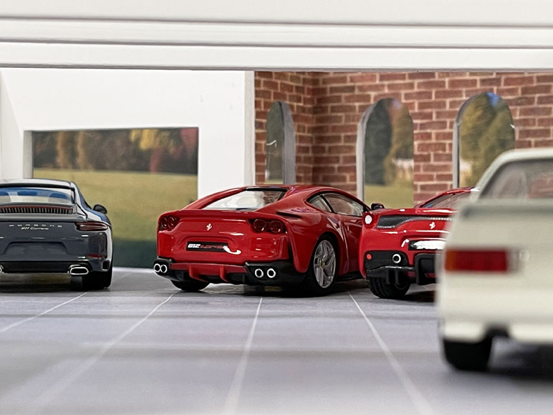 A small-scale car diorama featuring a detailed garage with four miniature luxury cars. Prominently, two red sports cars are parked side by side, facing outward. They are flanked by a gray and another white car. The backdrop resembles a brick wall with arched windows, reminiscent of elegant garden rooms. - a room in the garden