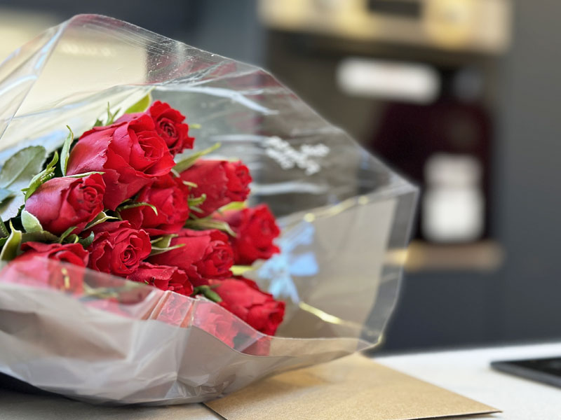 A bouquet of red roses wrapped in clear plastic is placed on a table. The background, reminiscent of serene garden rooms, is slightly blurred with indistinct objects, focusing the attention on the vibrant flowers. - a room in the garden