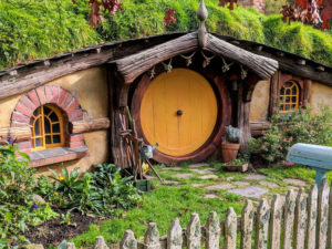 A picturesque, Hobbit-like house with a round yellow door set into a grassy hillside. The house has charming features including small arched windows, wooden beams, a stone pathway, plants, and a white picket fence in front. This quaint dwelling is the perfect Garden room BnB surrounded by lush greenery. - a room in the garden