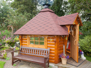 A charming wooden garden cabin, perfect as a Garden room BnB, boasts a hexagonal shingled roof adorned with string lights around the window and doorway. Surrounded by lush greenery and flowers, a wooden bench sits in front. The open door reveals the cozy interior awaiting guests. - a room in the garden