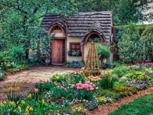 A charming, small cottage with a thatched roof and arched windows is nestled in a lush garden. The garden, belonging to this delightful Garden room BnB, is filled with vibrant flowers in various colors, including tulips and daffodils, along with neatly arranged greenery. A wooden trellis stands prominently in the foreground. - a room in the garden