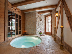 A cozy indoor spa room with rustic wooden beams and stone walls features a round, sunken hot tub. There's a large window overlooking the garden room, a wooden door leading outside, candles on antique holders, and neatly folded towels with flowers on the floor, evoking a serene atmosphere in this charming BnB. - a room in the garden