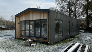A modern, rectangular tiny house with a blend of wood and dark metal siding embodies both style and durability. Large glass doors and windows dominate one side of the home. The surrounding yard is dusted with a light layer of snow, bare trees, and construction materials ready for upcoming maintenance. - a room in the garden
