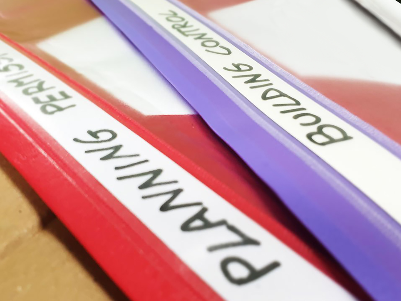 Close-up of three document binders, each labeled in black handwriting on white labels. The red binder is labeled "PLANNING," the purple binder is labeled "BUILDING CONTROL," and the stack beneath includes one marked "PLANNING PERMISSION" for a garden room. The binders are resting on a surface. - a room in the garden