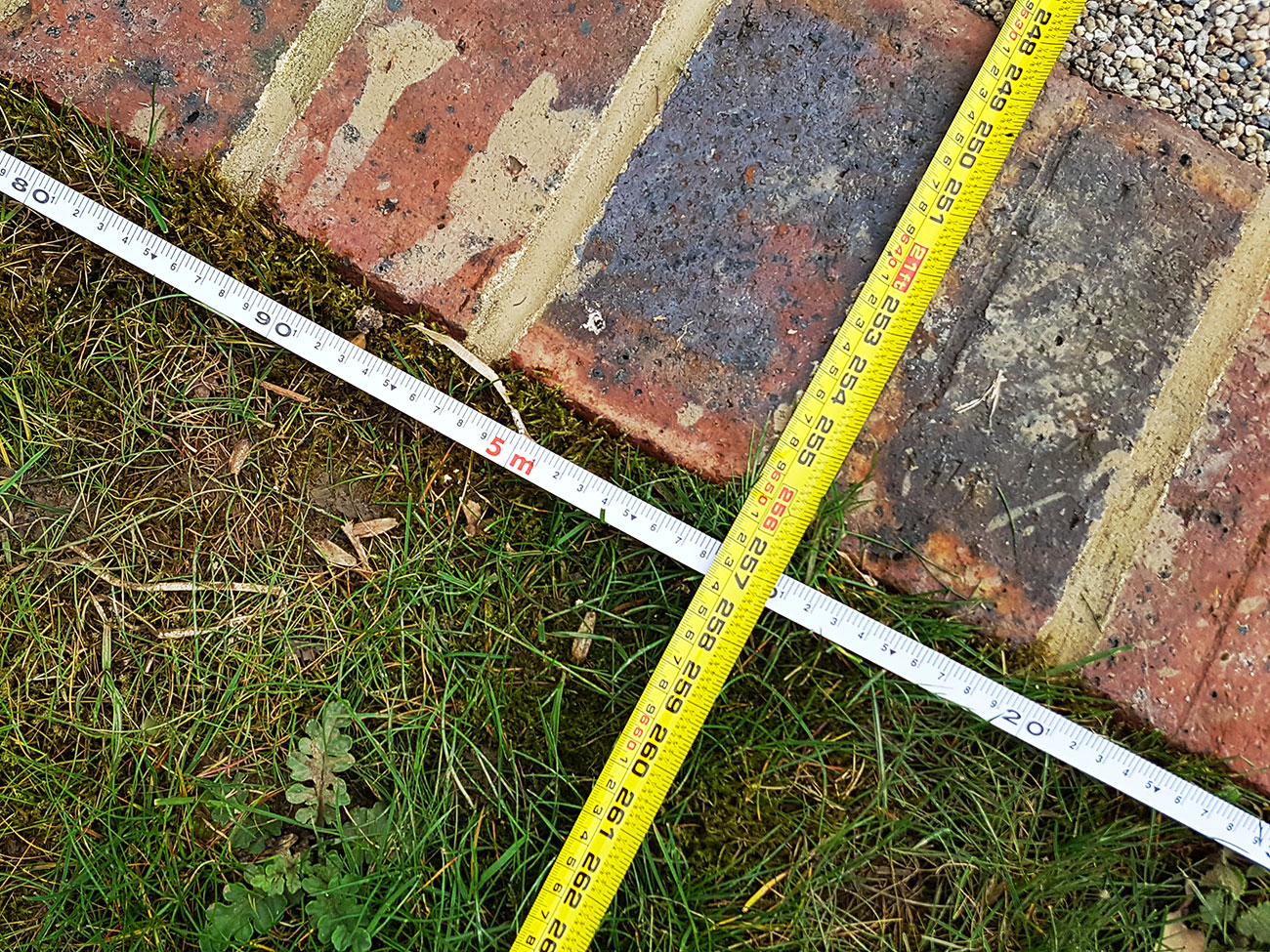 Two measuring tapes are arranged on the ground forming a cross, exemplifying meticulous architectural design. One is a white tape measure with meter markings, and the other is yellow with centimeter markings. They are placed over grassy soil and a brick-paved area. - a room in the garden