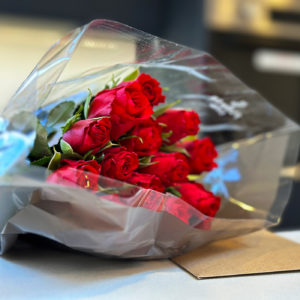 A bouquet of red roses wrapped in clear plastic lies on a surface. The flowers are fresh, with vibrant green stems and leaves about them. The background is slightly blurred, focusing attention on the roses. - a room in the garden