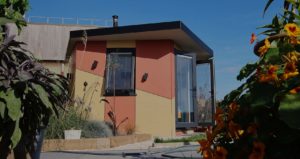 A small, modern house with angular design features in shades of red, yellow, and beige, surrounded by colorful plants and flowers. The house has large glass doors and windows, with a clear blue sky in the background. - a room in the garden