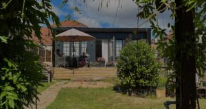 A backyard garden with a grassy path leads to a raised wooden deck with a table and chairs under an umbrella. Resembling serene garden rooms, two people sit at the table, surrounded by colorful flowers. A tree arch frames the image, and a house with open French doors is in the background. - a room in the garden