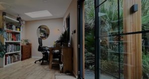 A modern home office with a large desk, ergonomic chair, computer, and monitor. A bookshelf filled with books is on the left, and a round mirror hangs on the back wall. Glass doors on the right open to lush greenery outside, blending this space seamlessly with garden rooms. The wooden floor adds a warm touch. - a room in the garden