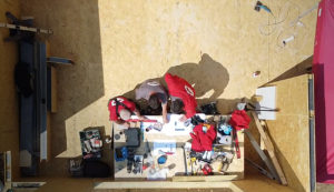 A top-down view of three people working together at a cluttered table. The table is scattered with tools, papers, and electronics. Two in red shirts and one in gray focus on a document amidst various items, as if strategizing the design for future garden rooms. - a room in the garden