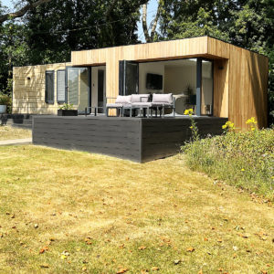 A modern, single-story house with a wooden exterior and a dark, raised deck featuring outdoor seating. The house has large glass doors that open up to the deck and garden rooms, surrounded by a grassy yard with some yellow flowers. Trees are visible in the background. - a room in the garden
