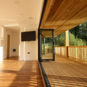 A modern, spacious room with wooden flooring and a wall-mounted TV, opening up to a large outdoor wooden deck with a railing. Sunlight streams through the deck, casting shadows on the floor. The area is surrounded by greenery. - a room in the garden