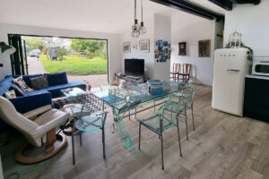 A modern living room and dining area with glass furniture. The space features a blue sectional sofa, a large open window with a view of a green yard, various wall art, a flat-screen TV, a vintage white refrigerator, and a wooden floor. - a room in the garden