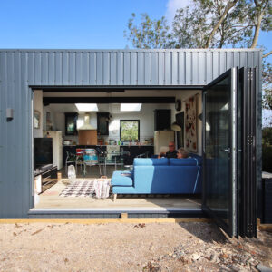 A modern, single-story container home with an open sliding glass door reveals the interior. Inside, a person is seen relaxing on a blue sofa. The home has a contemporary design with a kitchen and living area featuring sleek furniture and large windows. - a room in the garden