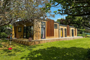 A modern, single-story wooden house with large glass doors and vertical wood slats. It is surrounded by green grass and trees, with a red hula hoop hanging on the left side of the deck and a colorful garden decoration nearby. The sky is clear and blue. - a room in the garden