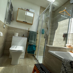 A modern bathroom featuring a toilet, a sink with a cabinet above, a glass-enclosed shower, and a bathtub. Towels hang on a wooden rack, and a small orange stool with black and white polka dots sits by the bathtub. The room is illuminated by natural light. - a room in the garden