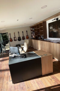 A well-lit office with wooden flooring, featuring a large wooden desk with a chair and computer. Behind the desk are a series of electric guitars mounted on the wall. A large bookshelf displays numerous items, and there's a piece of art on the opposite wall. - a room in the garden