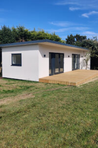 A modern, white, single-story house with a flat roof and large glass doors is situated on a grassy lawn. The house features a small wooden deck in front, and there is shrubbery and trees in the background under a clear blue sky. - a room in the garden