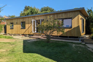 A modern wooden cabin with large windows and an open door stands in a sunny yard with lush green grass and a few small trees. The building features a simple, rectangular design and is partially shaded by taller trees in the background. - a room in the garden