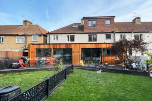 A two-story house with a wooden extension at the back features large glass windows. The backyard has a well-maintained lawn, a black fence, patio furniture with red, blue, and pink chairs, and various toys and gardening equipment scattered around. - a room in the garden