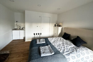 A cozy bedroom features a double bed with a patterned duvet and two neatly placed towels. The room has light-colored walls and wooden flooring. In the corner is a small kitchenette with a sink, stovetop, and mini-fridge. A white wardrobe stands next to the bed. - a room in the garden