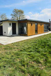 A modern, small building with a flat roof stands in a yard with green grass and a stone-paved path. The building features a mix of white plaster and natural wood siding with large windows. Trees and a blue sky with a few clouds are visible in the background. - a room in the garden