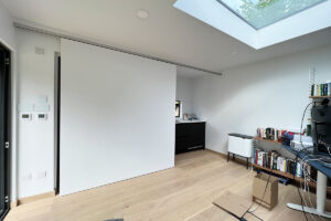 A minimalist room with white walls and light wooden flooring. There's a skylight in the ceiling, a sliding door on the left, bookshelves and a computer desk on the right, and a cabinet with a small plant in the back. Cardboard boxes are on the floor. - a room in the garden