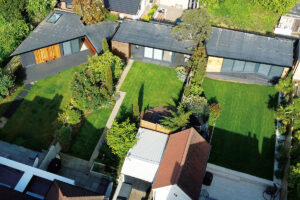 Aerial view of a residential area featuring four modern houses with flat roofs and large, neatly maintained grassy backyards. The houses are surrounded by greenery, including trees and bushes, and are separated by wooden fences and pathways. - a room in the garden