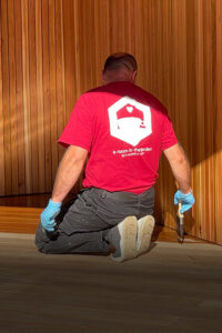 A person wearing a red shirt with a hexagon logo, kneels on the wooden floor, working with a tool on the wooden wall. The shirt has text that reads "a room in the garden... SET YOUR IMAGINATION FREE". They are wearing blue gloves and grey shoes. - a room in the garden