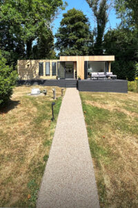 A modern tiny house with large windows and wood accents stands surrounded by lush greenery. A beige gravel pathway leads up to the house, which features an outdoor seating area on an elevated deck. The lawn is well-maintained with scattered trees and shrubs. - a room in the garden