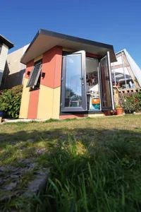 A small, modern, shed-like structure with colorful panels and large windows sits in a garden. The sky is blue and clear, and the ground in front of the structure is grassy. Various plants are growing around the building. - a room in the garden