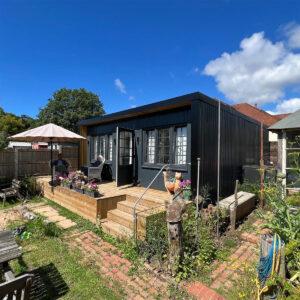 A modern shed, reminiscent of charming school buildings, with large windows and an open door sits on a wooden deck. Flowers in pots adorn the entrance. An outdoor seating area with an umbrella is to the left. The sky is clear with a few clouds. - a room in the garden